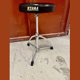 Černý bubenířský židle s kovovým chromovaným stojanem a třínožkou. Na sedáku je bílý nápis TAMA a pod ním text THE STRONGEST NAME IN DRUMS. Povrch se…