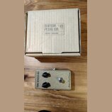 Kartonová krabice s nápisem BRITISH PEDAL CO. LONDON - ENGLAND. Pod ní je efekt pedál v béžovém provedení s kovovým spínacím tlačítkem a dvěma černým…