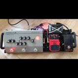 Sestava efektů na černém pedalboardu. Vlevo je stříbrný pedál s nápisy MODULE, TERRA-NOVA a BASS PREAMP PEDAL. Uprostřed je červený pedál s nápisy CO…