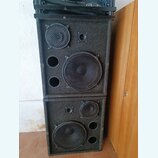 Dvě černé reproduktorové bedny s povrchem z tmavého kobercového materiálu, naskládané na sebe. Každá bedna obsahuje jeden velký a jeden malý reproduk…