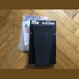 Černý volume pedál s textem M2 PASSIVE VOLUME. Na bocích jsou viditelné nápisy OUT a IN. Vedle pedálu je v plastovém sáčku dokumentace s nápisem MORL…