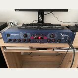 Modrý kytarový procesor s kovovým šasi a názvem V-AMP PRO a logem BEHRINGER. Na předním panelu jsou ovládací knofíky, červené tlačítka A až E a digit…