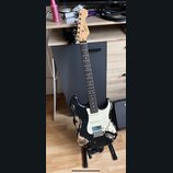 Černá elektrická kytara s bílým pickguardem a světle hnědým hmatníkem. Na hlavě je vidět nápis "Squier". Tělo nástroje vykazuje výrazné opotřebení v…