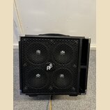 Černý basový reproduktor v kostce s povrchem z texturovaného vinylu a kovovými rohy. Přední stranu tvoří černá kovová mřížka s logem "pβ" v kruhu upr…