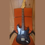 Černá elektrická kytara s tmelem v barvě přírodního dřeva. Na hlavě je viditelné logo Squier. Kytara má tři snímače, chromovaný most a ovládací knofl…
