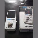 Bílý kytarový ladič s černým displejem a kovovým spínacím tlačítkem. Na přístroji jsou nápisy polytune mini, true bypass a tc electronic. Vedle ladič…