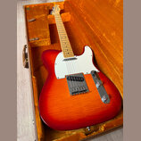 Elektrická kytara s červeno-oranžovým gradientem (sunburst) v hnědém kufru s oranžovou výplní. Nástroj má bílý pickguard, javorový hmatník a chromova…