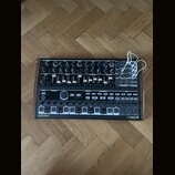 Černý analogový syntetizátor v dřevěném rámu s množstvím potenciometrů, posuvníků a tlačítek. Na spodním panelu jsou vlevo nápis ARTURIA a vpravo MIN…