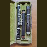 Tři části dřevěného dechového nástroje v tmavě hnědé barvě, umístěné v otevřeném kufru s zeleným polstrováním. Nástroj má kovové klapy a prstence v h…
