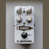 Bílý kytarový efekt s čtyřmi bílými oválnými knoflíky. Na pedálu jsou nápisy FAT CLEAN MID, LEVEL, TREBLE a logo SPARK BOOSTER s motivem zapálené záp…