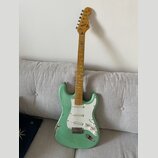 Světle zelená elektrická kytara s bílým pickguardem a javorovým hmatníkem. Na hlavě je viditelné logo Fender. Lak na těle vykazuje výrazné opotřebení…