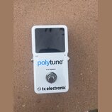 Bílý kytarový ladič s černým displejem. Na přední straně je modrý nápis polytune®, pod ním text true bypass a ve spodní části logo tc electronic®. Na…
