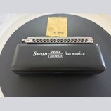 Stříbrná chromatická harmonika s kovovým povrchem a řadou otvorů. Vedle ní je černý plastový obal s bílým nápisem Swan 1664 CHROMATIC Harmonica. V po…