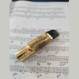 Zlatý kovový mouthpiece s černým zakončením v horní části. Povrch je viditelně poškrábaný a opotřebovaný. Na boku jsou vyryty nečitelné znaky. Nástro…