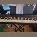 Černý digitální klavír značky YAMAHA umístěný na černém X-kovovém stojanu. Na klavíru je nápis YAMAHA a v zadní části je umístěna kniha s nápisem DIG…