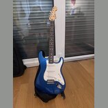 Modrá elektrická kytara s bílým pickguardem a javorovým hmatníkem. Na hlavě je viditelné logo Fender. Kytara stojí na černém stojanu a vedle ní je če…