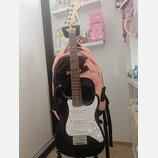 Černá elektrická kytara s bílým pickguardem a světle hnědým hmatníkem. Na hlavě nástroje je viditelné logo Squier. Kytara stojí na stojanu před růžov…