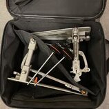V černé přepravní tašce jsou umístěny kovové části bicího pedálu v stříbrném provedení. Jsou zde vidět dvě nožní základny, spojovací tyče a dva černé…