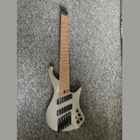 Stříbrná elektrická basa s šesti strunami a hnědým hmatníkem. Na korpusu je viditelné logo Ibanez. Nástroj má černý hardware a tři ovládací knoflíky…