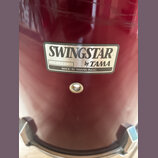 Detail červeného bubnu s nalepeným stříbrným štítkem. Na štítku je uveden nápis SWINGSTAR by TAMA a text MADE IN TAIWAN R.O.C. V levé části štítku je…