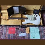 Bílá elektrická kytara typu Stratocaster s bílým pickguardem, umístěná v černém kufru. Na hlavě kytary je viditelné logo Fender. K nástroji patří cer…