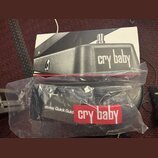 Fotografie zobrazuje krabici a plastový obal s manuálem k wah-wah pedálu. Na krabici je černobílá fotografie přístroje s nápisem cry baby. V plastové…