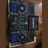 Černý DJ kontrolér Denon DJ Prime 4 s integrovaným displejem, na kterém je nápis engine dj a verze 5.0. Zařízení má dva jog-kola s modrým podsvícením…