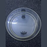 Dvě průhledné bubny na bicí nástroj v různých velikostech, položené na šedém povrchu. Na menším bubnu je v dolní části logo REMO a nápisy AMBASSADOR,…