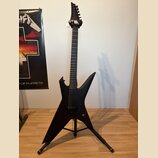 Černá elektrická kytara s ostře hranatým korpusem a špičatou hlavou, umístěná na černém stojanu. Nástroj má dva humbuckery a jeden ovládací knoflík.…