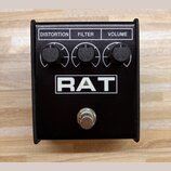 Černý efekt pedál s nápisem RAT v bílém obdélníku. V horní části jsou tři černé otočné ovladače s popisky DISTORTION, FILTER a VOLUME. Ve spodní část…