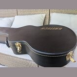 Černý tvrdý kufřík na kytaru s texturovaným povrchem a bílým prošíváním po okrajích. Na horní části je zlatý nápis GRETSCH. Kufřík má zlaté kovové zá…
