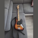 Elektrická basová kytara s korpusem v barevném přechodu od žluté přes oranžovou k červené a černé. Nástroj má černý pickguard, stříbrný most a ladící…