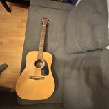 Akustická kytara s cutawayem v přirozené dřevěné barvě. Na hmatníku je vidět logo "Fender". Nástroj leží na šedé pohovce.