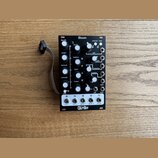 Černý modulární syntetizátor s bílými popisky a ovládacími prvky. Na horní části je nápis Bloom, v dolní části bílý panel s logem QU-Bit a výstupy oz…