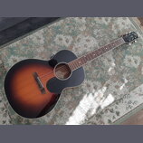 Akustická kytara s vyřezem v těle a tmavým, přechodovým lakem (sunburst) od černé k hnědé. Na hlavě je viditelné logo "Epiphone". Uvnitř otvoru kytar…