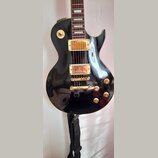 Černá elektrická kytara s gold hardwarem a hnědým hmatníkem. Na těle jsou dva humbuckery, čtyři otočné knofíky a přepínač s nápisy RHYTHM a TREBLE. P…