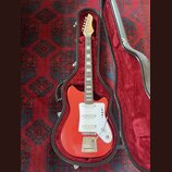 Červená elektrická kytara v černém tvrdém pouzdře s červeným vnitřkem. Nástroj má bílý pickguard, tři snímače a chromovaný most s tremolem. Na hlavě…