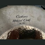 Detailní záběr na povrch činchy z kovu. Na povrchu je černý nápis „Custom Hybrid Crash“ a pod ním označení „19"/48cm“. Levá část fotky ukazuje kovaný…