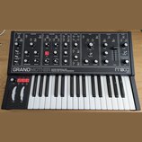 Černý semi-modulární analogový syntetizátor s bílými klávesami. Na předním panelu je nápis GRANDMOTHER SEMI-MODULAR ANALOG SYNTHESIZER a logo moog. P…