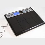 Černý elektronický bicí pad s osmi gumovými polštářky. V horní části je stříbrný panel s modrým displejem, ovládacím knoflíkem a řadou tlačítek. Na p…