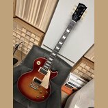 Elektrická kytara s tónem sunburst, bílým pickguardem a trapezoidovými vložkami na hmatníku. Na hlavě je vidět nápis Gibson, model Standard a podpis…