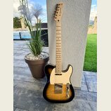 Elektrická kytara s tělem v provedení sunburst a bílým pickguardem. Na hlavě hředu je viditelné logo Fender. Kytara má světle dřevěný hmatník a zlaté…