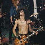 Fotografie zachicuje hudebníky během koncertu. V popředí je muž s kytarou v přirozeném dřevěném provedení a zlatými hardwarem. V pozadí je vidět část…