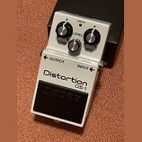Bílý efekt pedál s nápisy Distortion a DS-1. Na horní části jsou tři ovládací knoflíky s popisky LEVEL, TONE a DIST a červená kontrolka s nápisem CHE…
