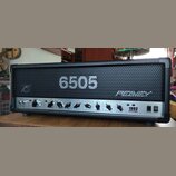 Kytarový zesilovač s černým povrchem a kovovou mřížkou. Na mřížce je bílý nápis 6505 a logo PEAVEY. Spodní ovládací panel obsahuje různé potenciometr…