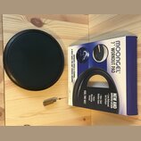 Černý tréninkový pad na bubny s kulatým tvarem. Vedle něj je modrá krabice s nápisy MOONGEL®, 7" WORKOUT PAD, FEEL THE GEL!, IMPROVES SPEED & STAMINA…