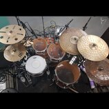 Pohled shora na bicí sadu s několika činely a mikrofony. Na bílém bedenku je viditelné logo Mapex, stejné logo je i na dvou tom-tamech v hnědém prove…