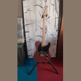 Elektrická kytara s růžovým tělem a černým pickguardem umístěná na stojanu. Na hlavě hředu je vidět nápis Fender TELECASTER. Hmatovnice a krk jsou vy…