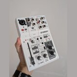 Bílá mixovací konzole Yamaha AG03. Na horní části jsou vstupy pro mikrofon, kytaru a linkové vstupy s nápisy „GUITAR“, „MONO“ a „LINE“. Přední panel…