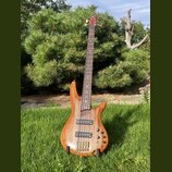 Dřevěná basová kytara s přírodním povrchem a hnědým hmatníkem. Na těle jsou dva černý humbuckery, čtyři ovládací knofíky a čtyři ladicí struny u most…